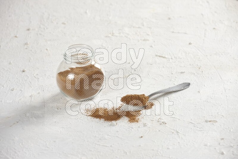 Herbs glass jar full of cinnamon powder with a metal spoon full of powder on a textured white background