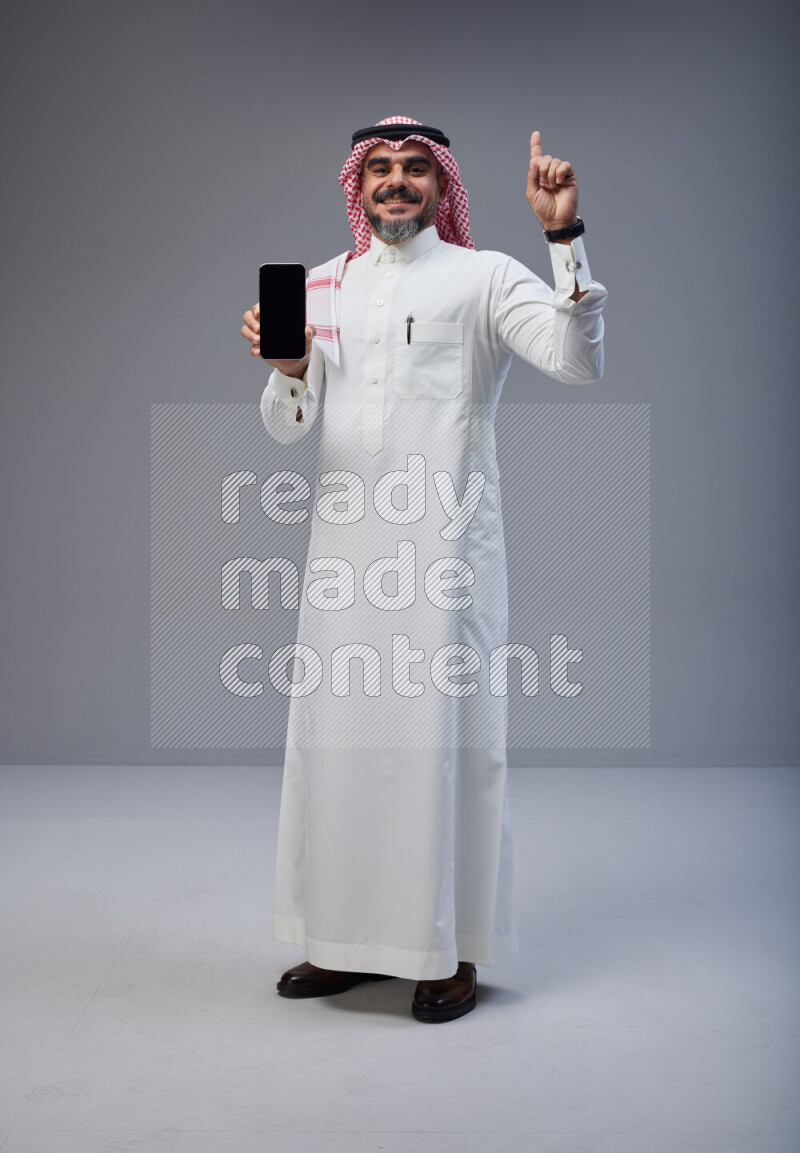 Saudi man Wearing Thob and red Shomag standing showing phone to camera on Gray background