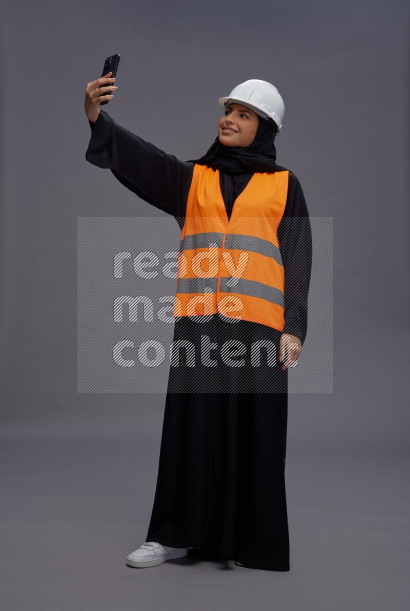 Saudi woman wearing Abaya with engineer vest standing taking selfie on gray background