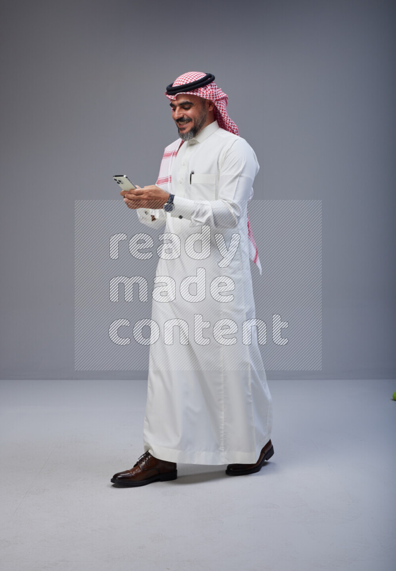 Saudi man Wearing Thob and red Shomag standing texting on phone on Gray background