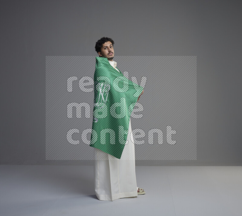 A Saudi man standing wearing thob wrapping big Saudi flag on gray background