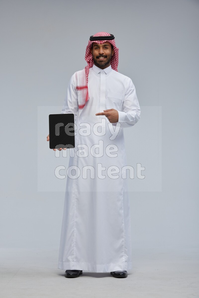 Saudi man Wearing Thob and shomag standing showing tablet to camera on Gray background