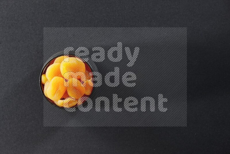 A black pottery bowl full of dried apricots on a black background in different angles