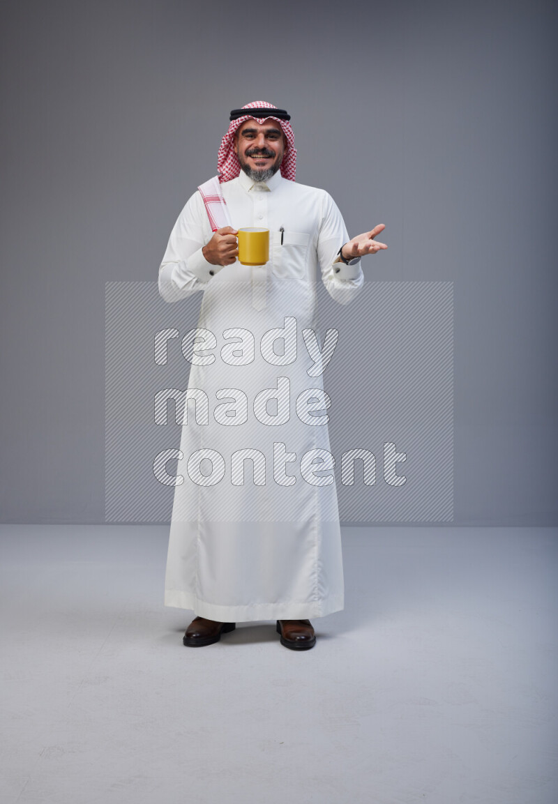 Saudi man Wearing Thob and red Shomag standing holding mug on Gray background