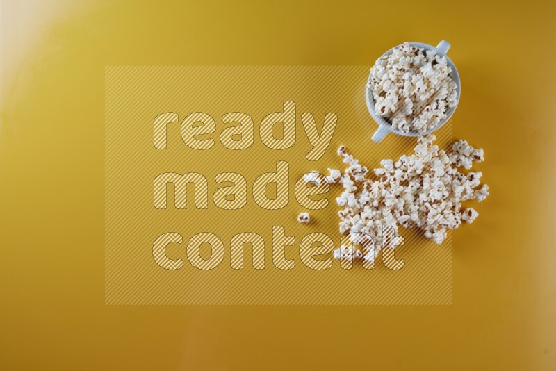 A white ceramic bowl full of popcorn with popcorn beside it on a yellow background in a top view shot