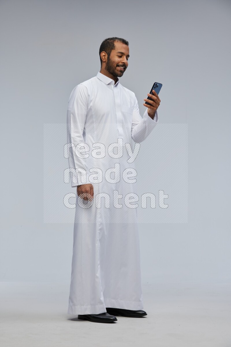 Saudi man wearing Thob standing texting on phone on Gray background