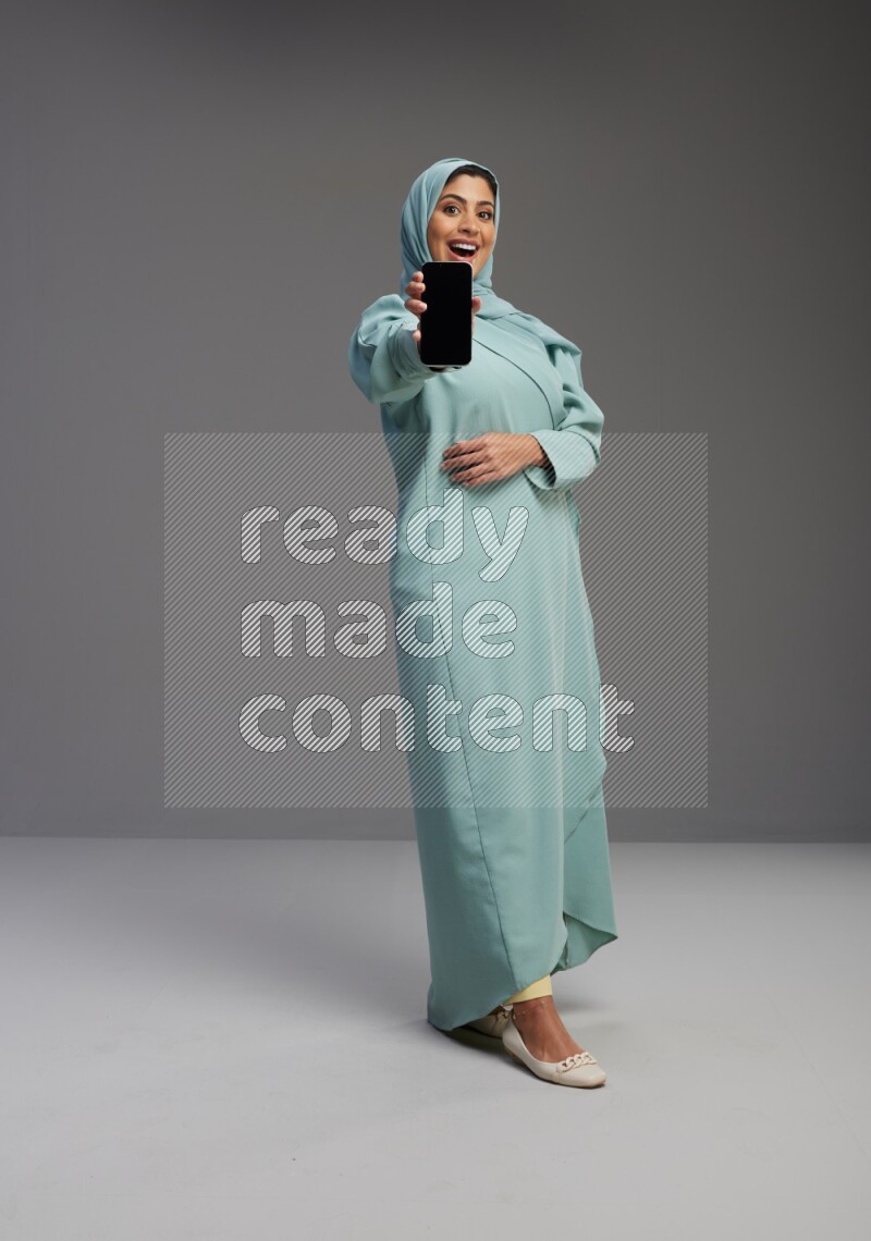 Saudi Woman wearing Abaya standing showing phone to camera on Gray background