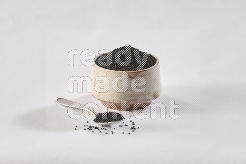 A beige pottery bowl and a metal spoon full of black seeds and more seeds spread on a white flooring