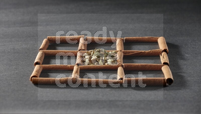 9 squares of cinnamon sticks full of cardamom in the middle surrounded by nutmeg, cinnamon, bay laurel leaves, cloves, cumin, dried ginger, dried basil and star anise on black flooring