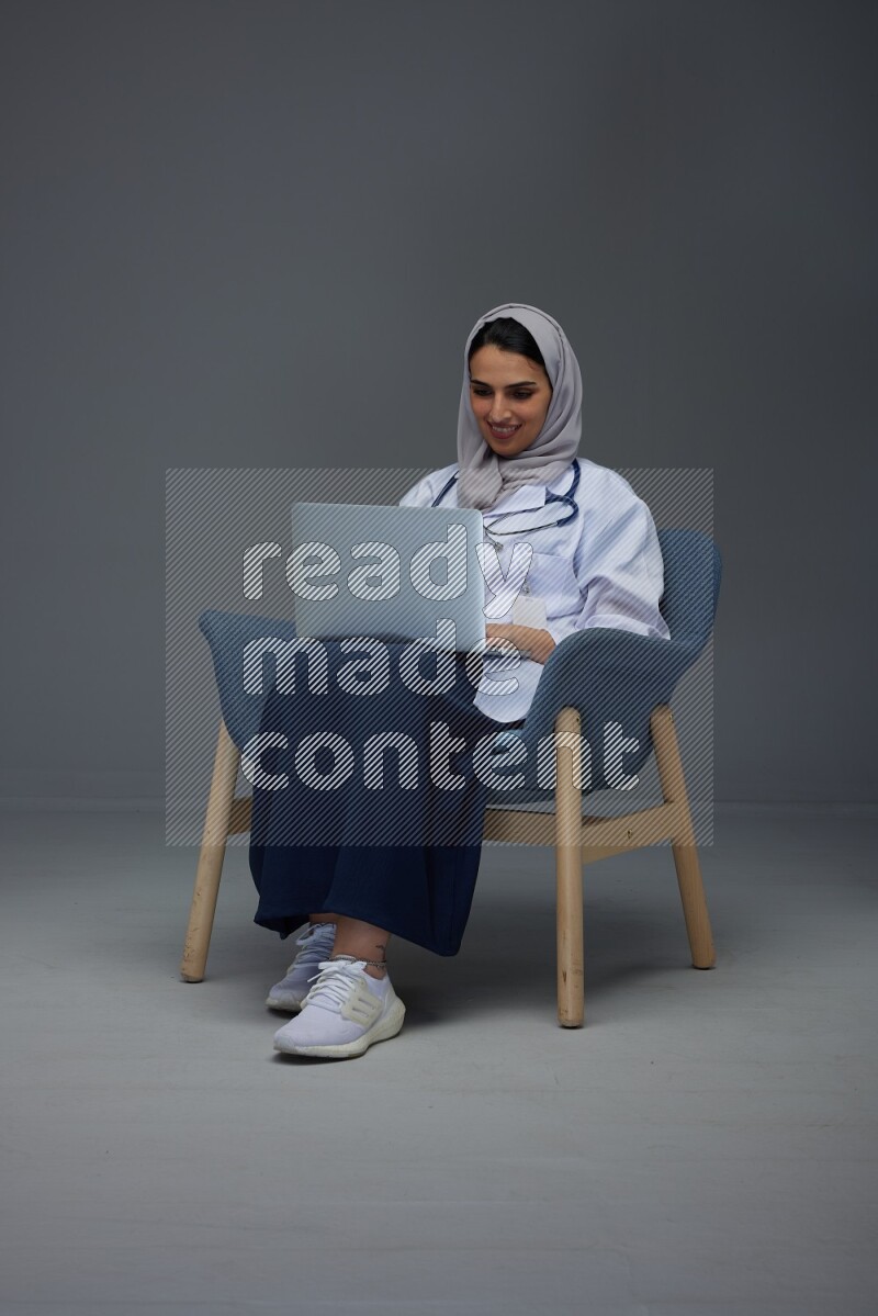 A doctor wearing a light gray head scarf sitting on a dark grey chair and using a laptop eye level on a grey background