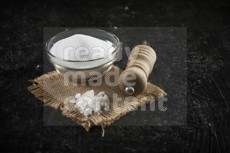 A glass bowl full of white salt with a wooden grinder on a burlap fabric all on black background