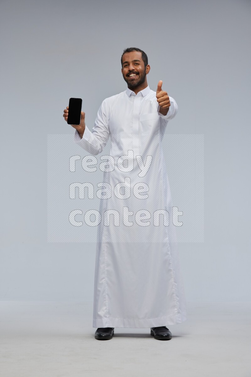 Saudi man wearing Thob standing showing phone to camera on Gray background