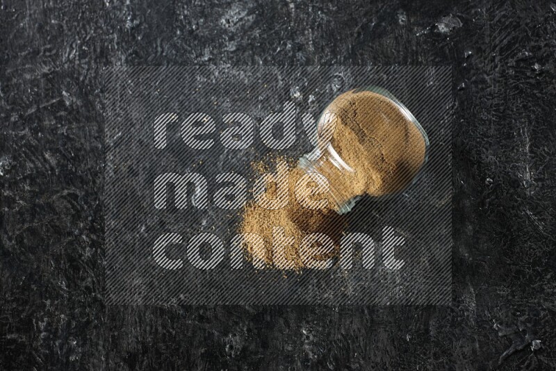 A flipped glass spice jar full of cumin powder and powder spilled out on a textured black flooring