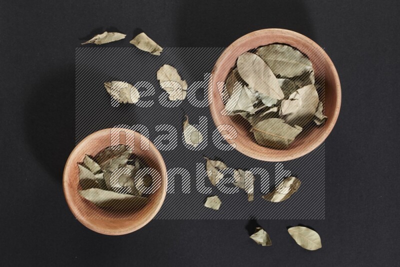 2 wooden bowls full of dried bay leaves with more leaves spread on black flooring