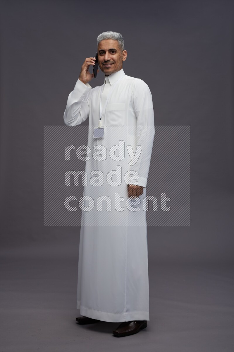 Saudi man wearing thob with neck strap employee badge standing talking on phone on gray background