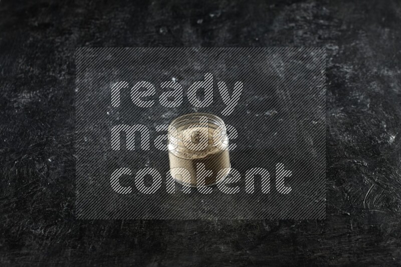 A glass jar full of cardamom powder on textured black flooring