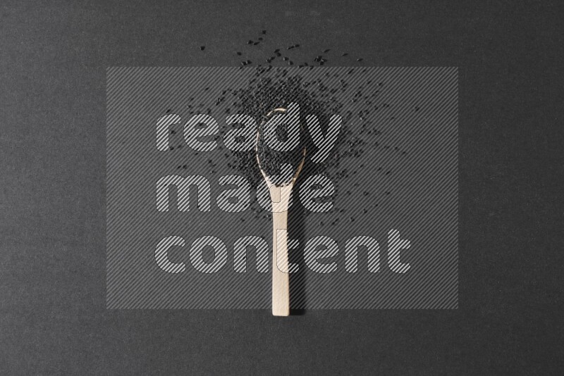 A wooden spoon full of black seeds and surrounded by seeds on a black flooring