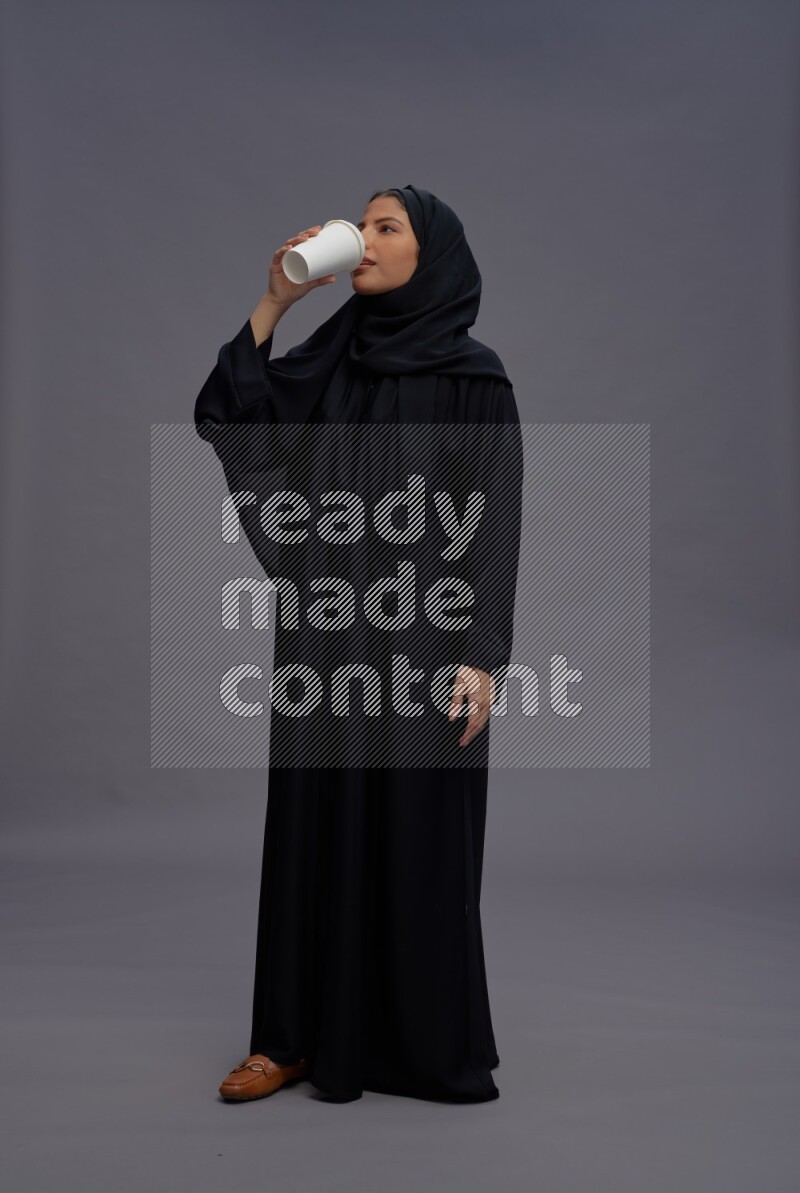 Saudi woman wearing Abaya standing holding paper cup on gray background