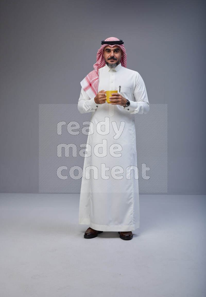 Saudi man Wearing Thob and red Shomag standing holding mug on Gray background