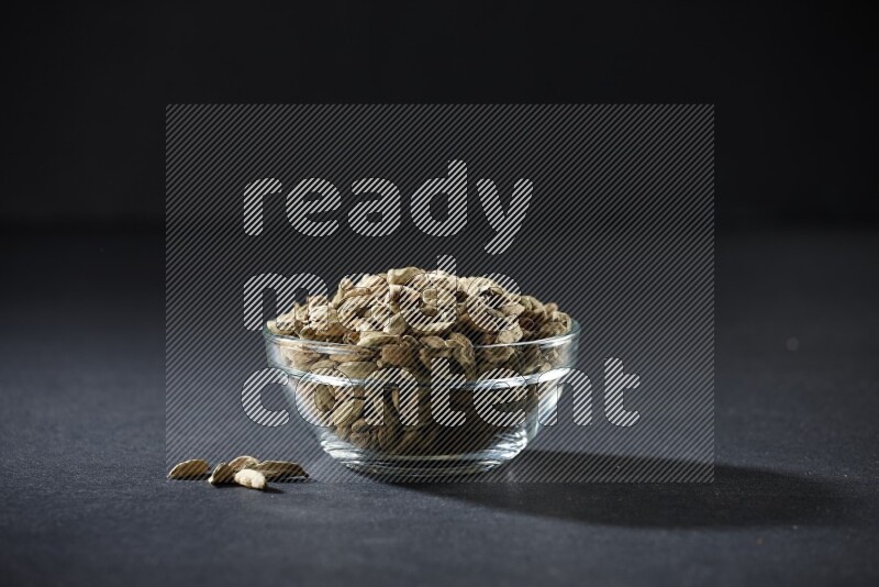 A glass bowl full of cardamom and more seeds spreaded beneath the bowl on black flooring