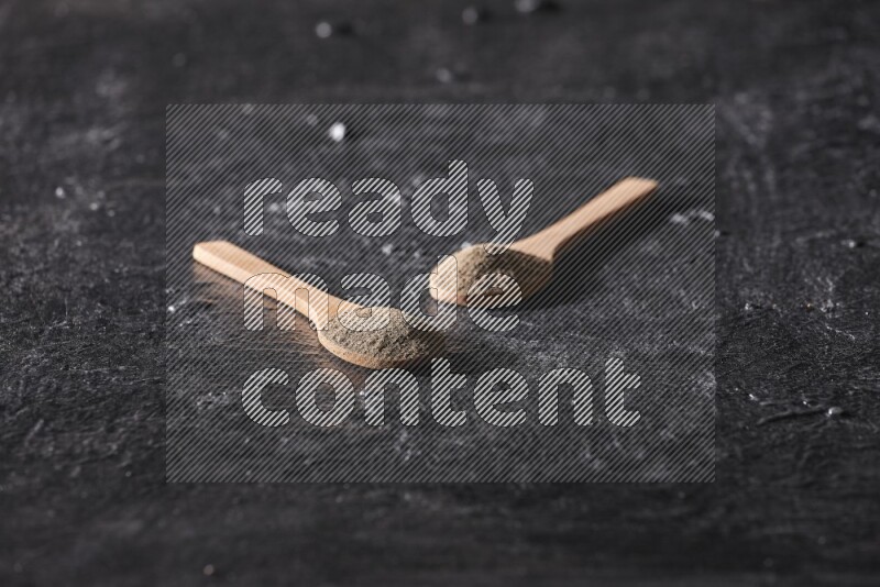 2 wooden spoons full of black pepper powder on a textured black flooring