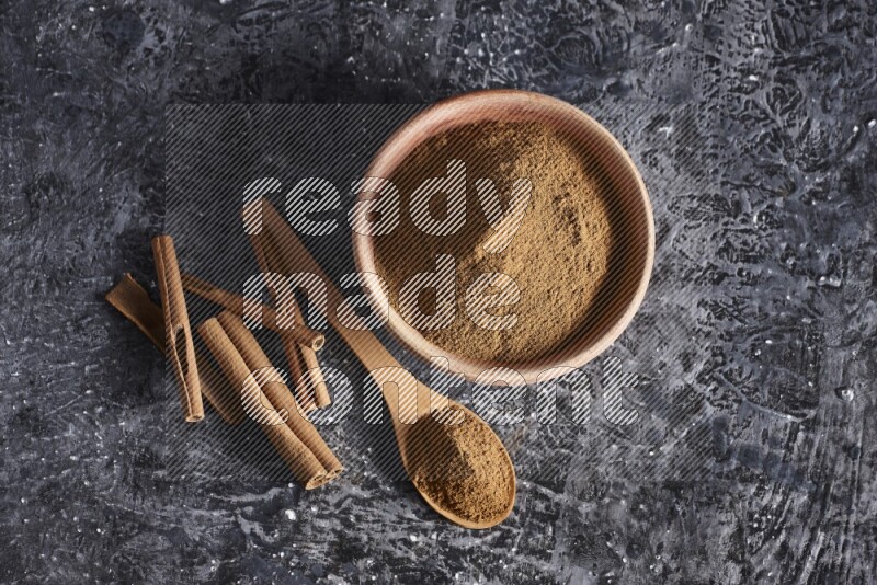 wooden bowl full of cinnamon powder and a wooden spoon full of it with cinnamon sticks on a textured black background