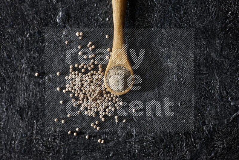 A wooden spoon full of white pepper powder with white pepper beads on textured black flooring