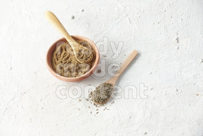 A wooden bowl and 2 wooden spoons full of cumin powder and cumin seeds on textured white flooring