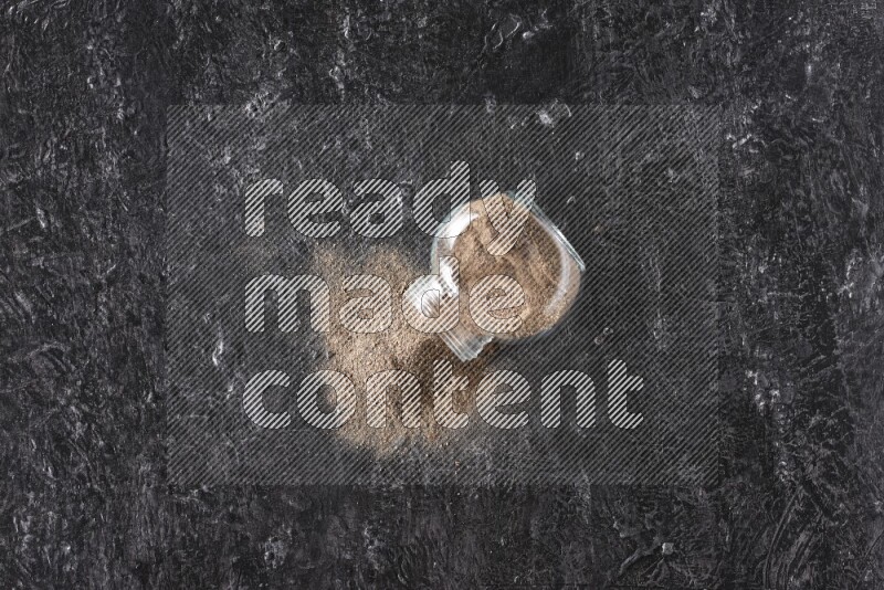 A flipped glass spice jar full of black pepper powder on textured black flooring