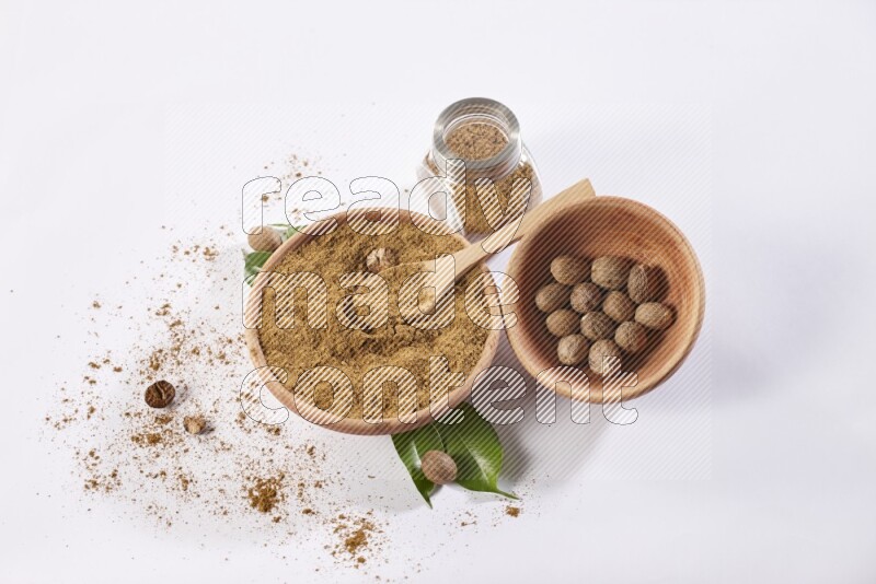 وعاء خشبي به ملعقة مع وعاء بهارات زجاجي جميعها ممتلئة ببودرة جوزة الطيب ووعاء خشبي ممتلئ بحبوب جوزة الطيب الكاملة على خلفية بيضاء