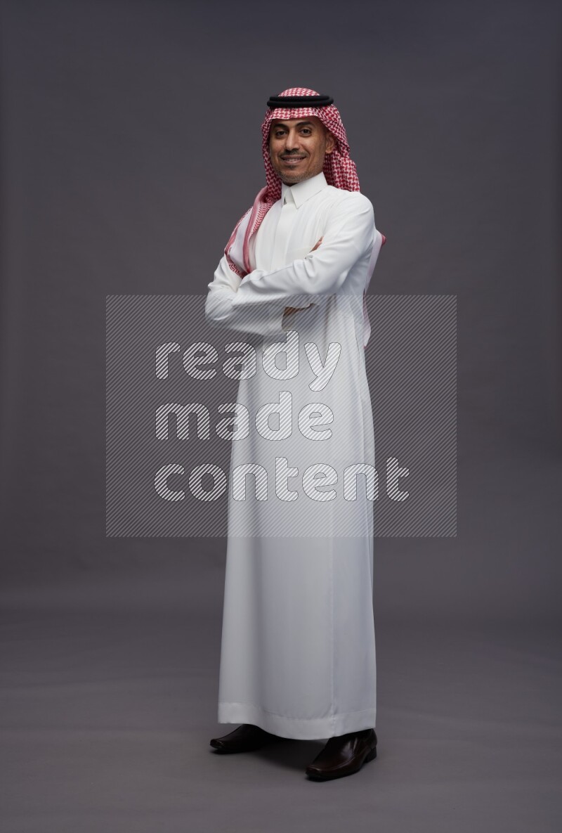 Saudi man wearing thob and shomag standing with crossed arms on gray background