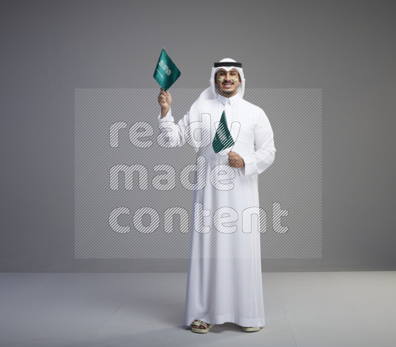 A Saudi man standing wearing thob and white shomag with face painting raising small saudi flag on gray background