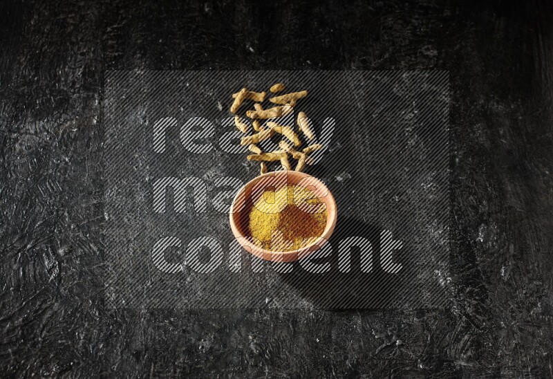 A wooden bowl full of turmeric powder with dried turmeric fingers on textured black flooring