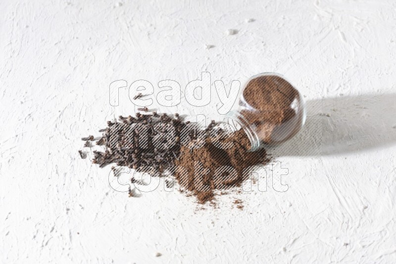 A glass spice jar full of cloves powder flipped and powder came out of it with cloves spread on textured white flooring