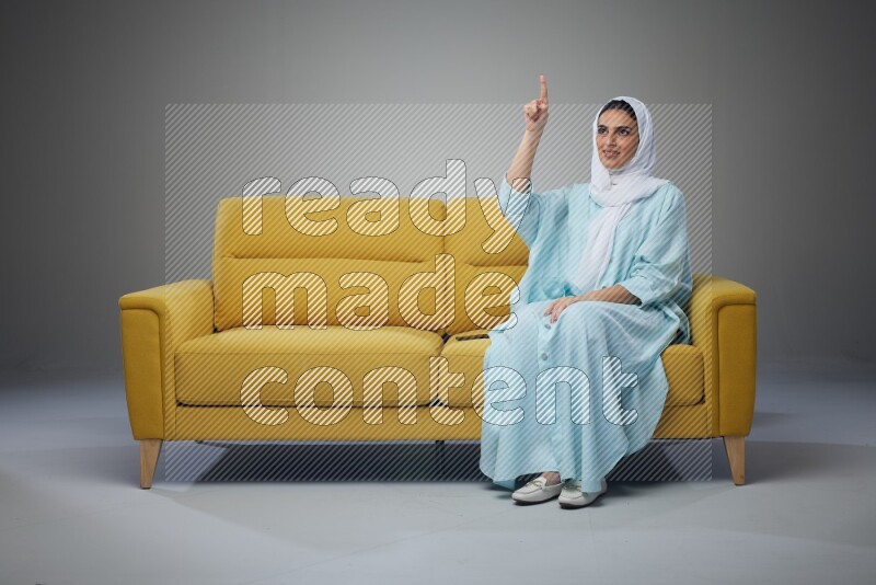 A Saudi woman wearing a light blue Abaya and white head scarf sitting on a yellow sofa and thinking and pointing up eye level on a grey background