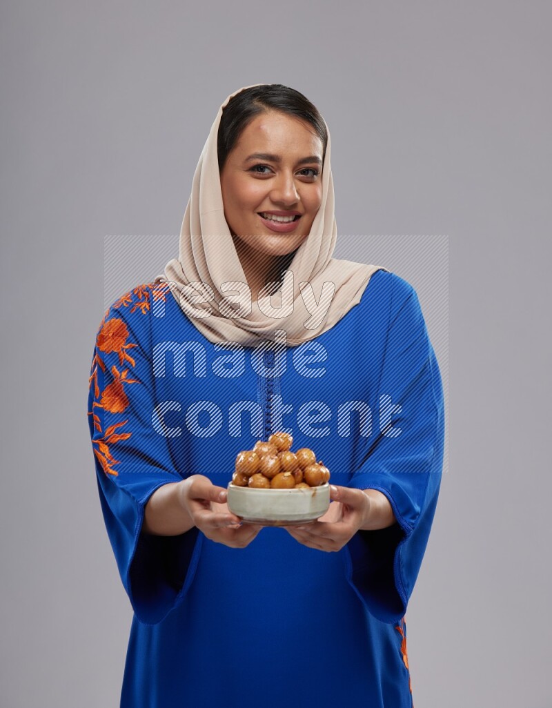 A Saudi woman standing wearing Jalabeya holding a plate of luqaimat