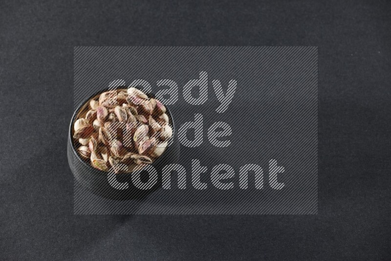 A black pottery bowl full of peeled pistachios on a black background in different angles