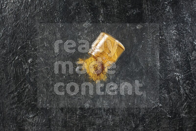 A flipped glass jar full of turmeric powder and powder spilled out of it on a textured black flooring