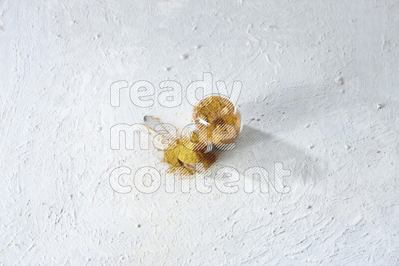 A flipped glass spice jar and a metal spoon full of turmeric powder and powder spilled out of it on textured white flooring