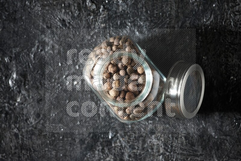 A glass spice jar full of allspice whole balls on a textured black flooring