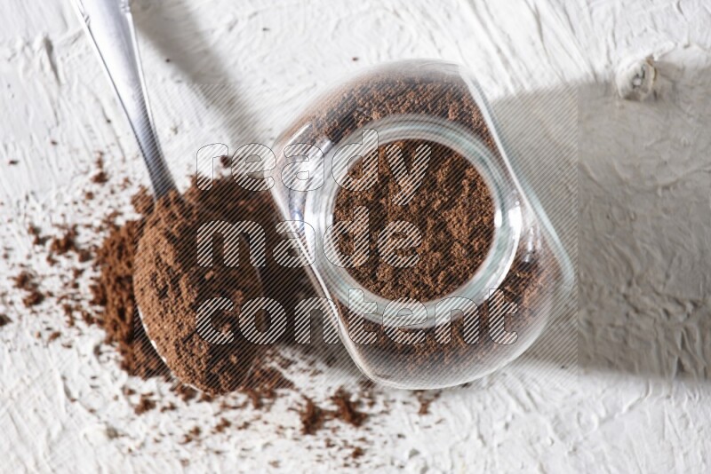 A glass spice jar and a metal spoon full of cloves powder on textured white flooring