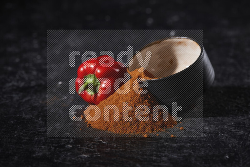 A black pottery bowl full of ground paprika powder with fallen powder from it on black background