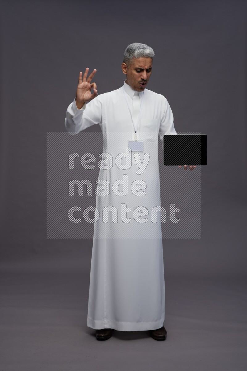 Saudi man wearing thob with neck strap employee badge standing showing tablet to camera on gray background