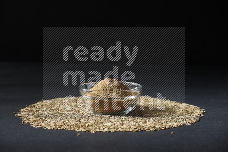 A glass bowl full of cumin powder surrounded by cumin seeds on black flooring
