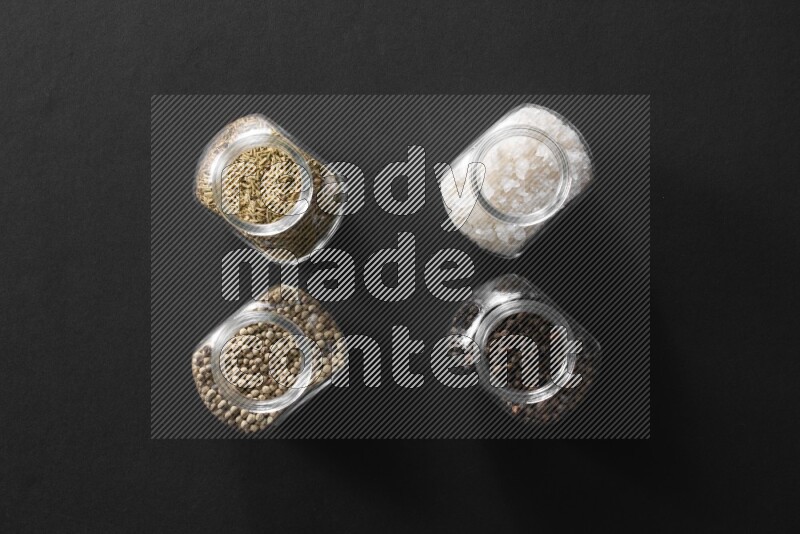4 glass spice jars full of salt, black peppers, white peppers and cumin on black flooring