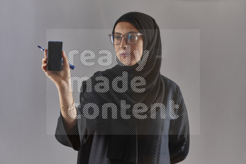 Woman in black abaya, hijab and glasses holding a marker pen to write on a transparent board with different reactions and expressions on grey background