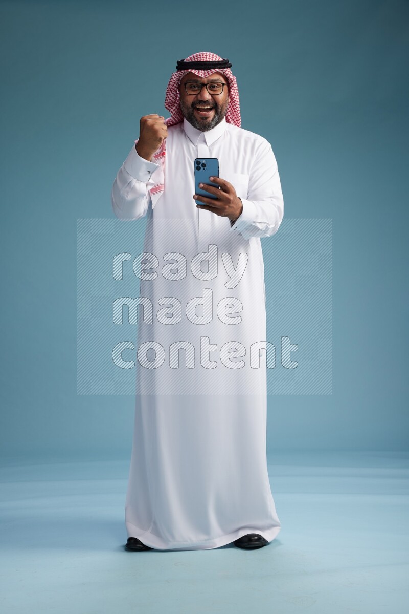 Saudi Man with shimag Standing texting on phone on blue background
