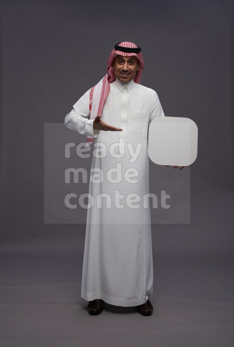 Saudi man wearing thob and shomag standing holding social media sign on gray background