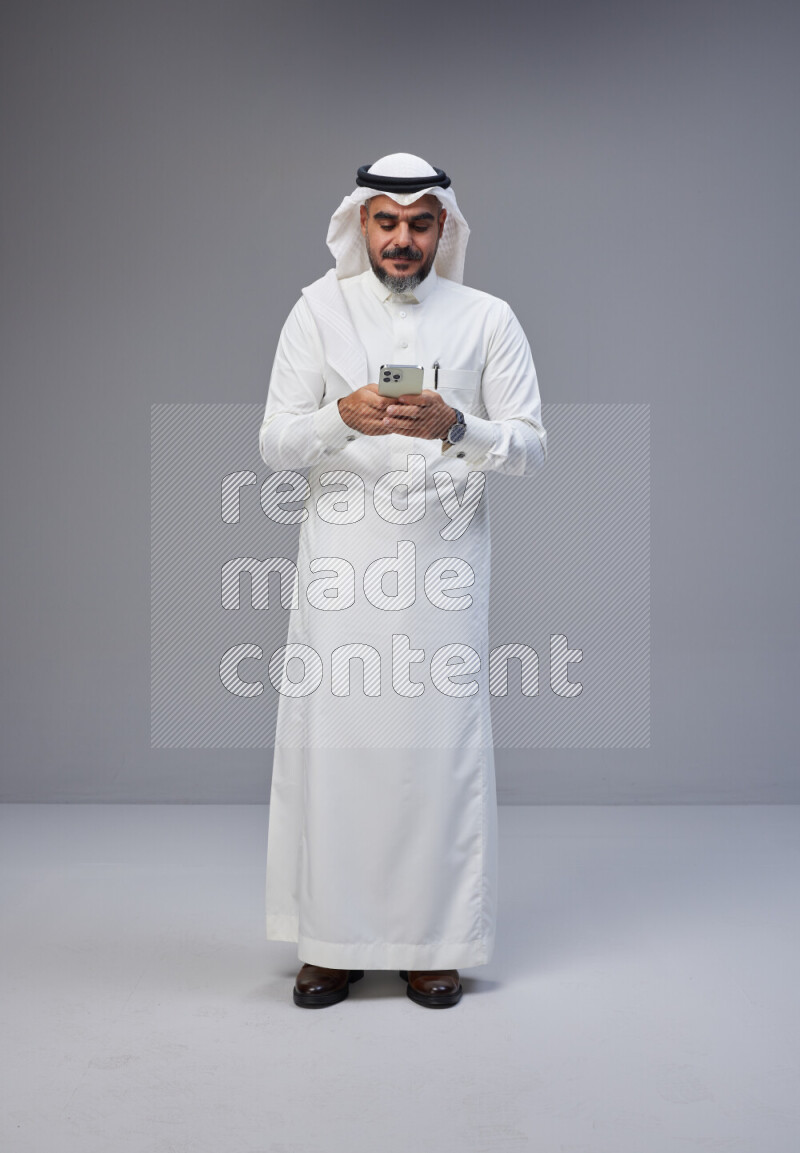 Saudi man Wearing Thob and white Shomag standing texting on phone on Gray background