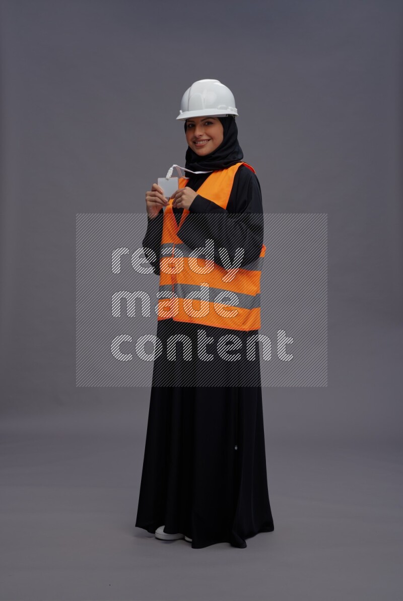 Saudi woman wearing Abaya with engineer vest with neck strap employee badge standing interacting with the camera on gray background